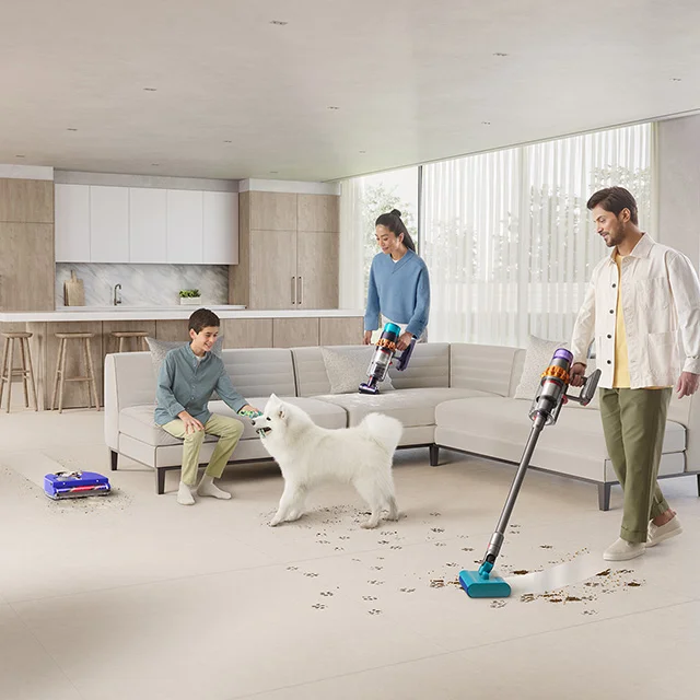 A family living area with child and dog, a robot vacuum, plus a parent using handheld cordless vacuum