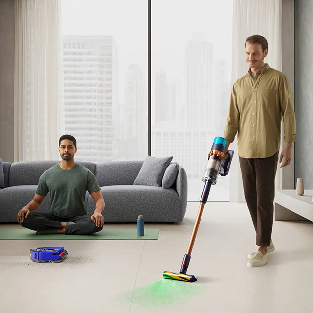 A man meditating on a yoga mat while robot vacuum cleans around him, with a man cleaning the floor with a cord free vacuum