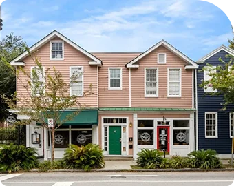 Two-story pink storefront with green door and white trim, flanked by trees and neighboring buildings.