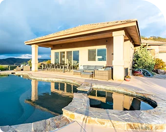 Luxury home with covered patio and stone-edged pool overlooking scenic hills under a cloudy sky.