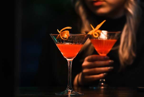 A woman drinking a pink cocktail from a martini glass, garnished with a twist of orange peel.