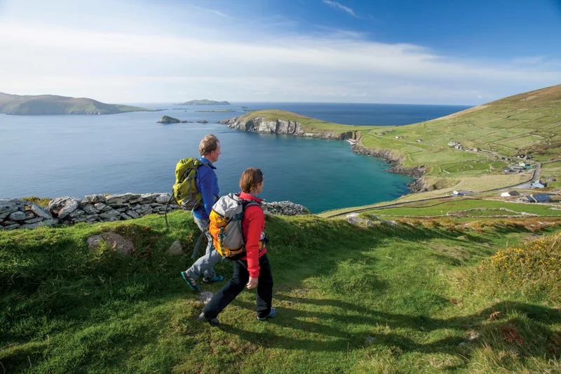 A man and a woman in hiking gear walk along a green hillside overlooking the ocean.