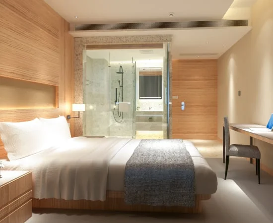Image description: Hotel room with a white bed and light natural wood furnishings with a clear glass bathroom in the room.