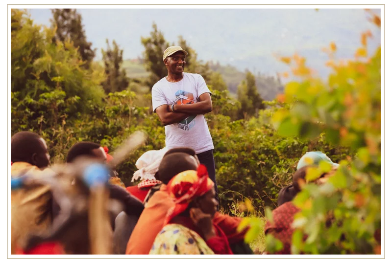 David Rubanzangabo stands outdoors at Huye Mountain with arms crossed, smiling as a group of people sit nearby, surrounded by lush greenery and a hillside landscape.