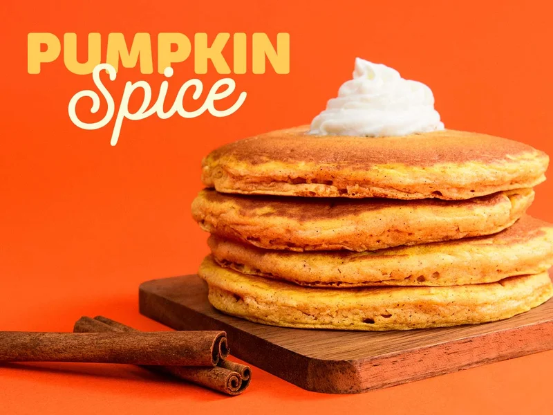 A Full Stack of Pumpkin Spice Pancakes with whipped topping on a wood cutting board, with two sticks of cinnamon beside it, against an orange background.