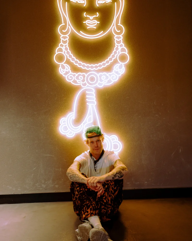 Person sitting on the floor in front of a wall with a glowing neon outline of a figure's face and necklace. The person is wearing a green cap, light-colored shirt, and patterned pants, with their arms resting on their knees and looking directly at the camera.