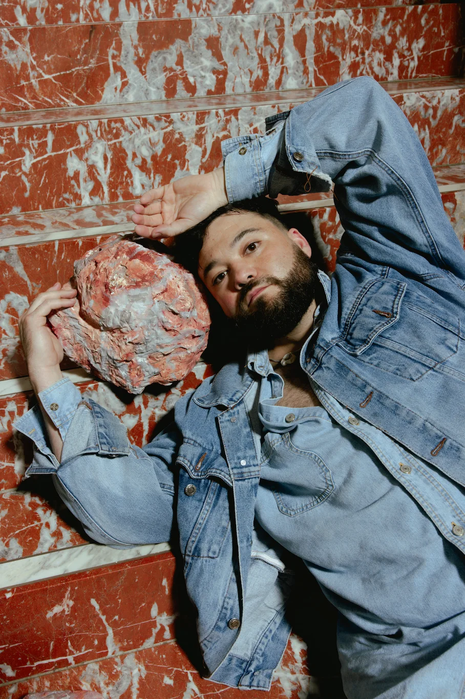 "Person lying on red and white marbled stairs, dressed in denim, with a textured object in hand. The person appears relaxed, with one arm resting on their head and the other holding the object close to their face."