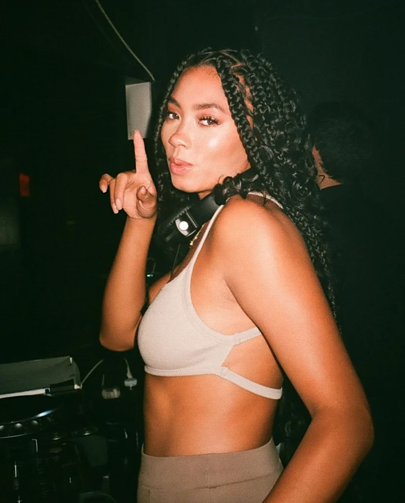 Person wearing a beige top, posing confidently with a playful expression while holding one finger up. The scene takes place at Evangeline in Ace Hotel Toronto, with a set of headphones around their neck."