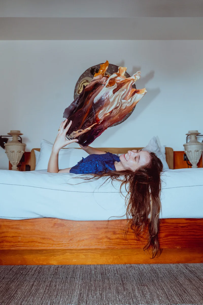 Person lying on a bed, smiling and holding up a large, abstract glass sculpture with swirls of warm colors. The room features decorative elements, including vases on either side of the bed, adding to the artistic and serene atmosphere.
