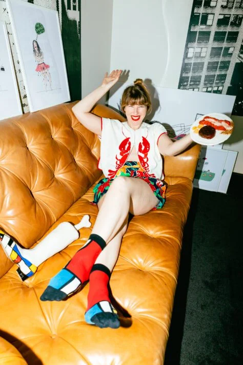 Person sitting on a tan leather couch, smiling and raising one hand while holding a plate with food in the other. They are wearing a colorful outfit with a lobster design on the shirt and vibrant socks. The room has an artistic vibe, with framed artwork leaning against the wall.