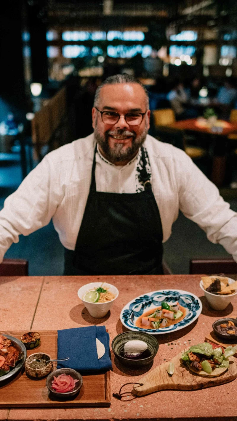 Chef smiling while standing behind a table filled with a variety of plated dishes. The table is set with a mix of vibrant, artfully presented foods, reflecting the inviting ambiance of the restaurant.