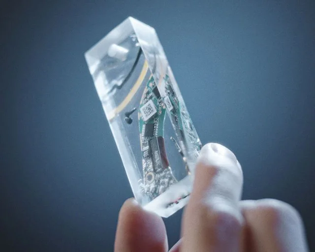 A hand holds the tech slice, a small cuboid of clear resin that contains parts relating to the Dyson OnTrac headphones