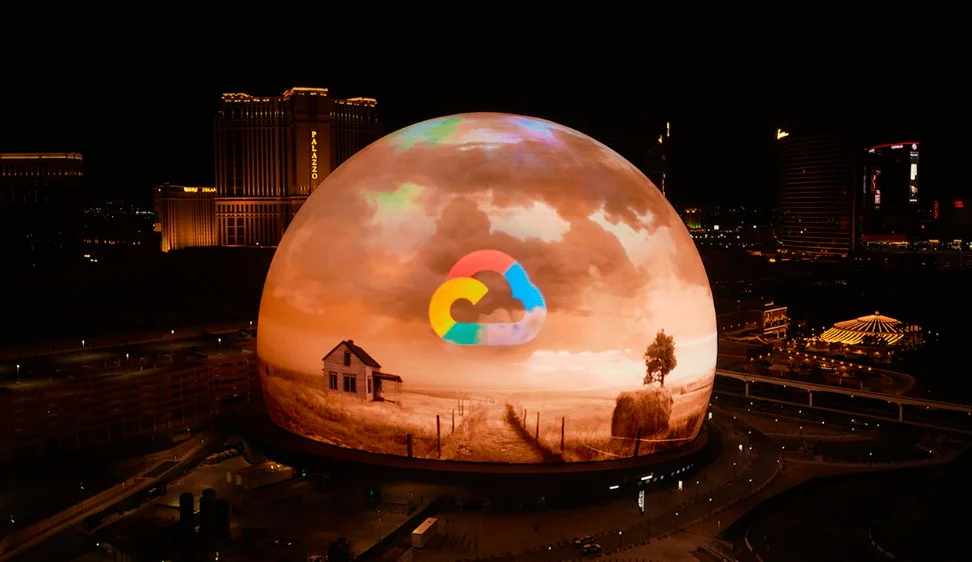 Night aerial view of the Las Vegas Sphere displaying a rainbow Google Cloud icon and a scene from the Wizard of Oz.