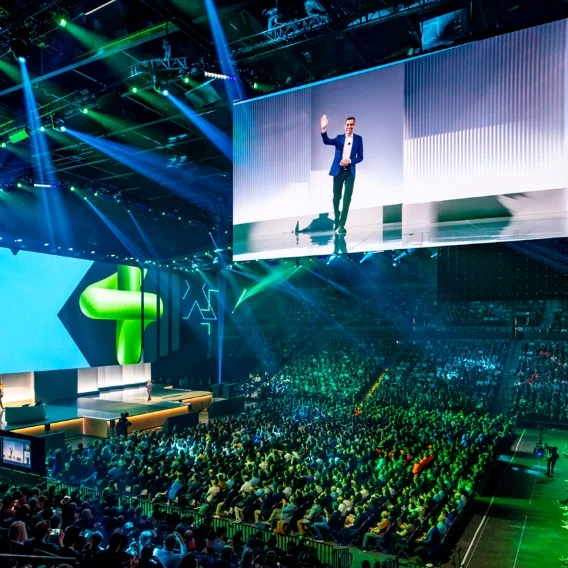 Large indoor event with a speaker on screen above a packed audience, illuminated by green and blue spotlights.