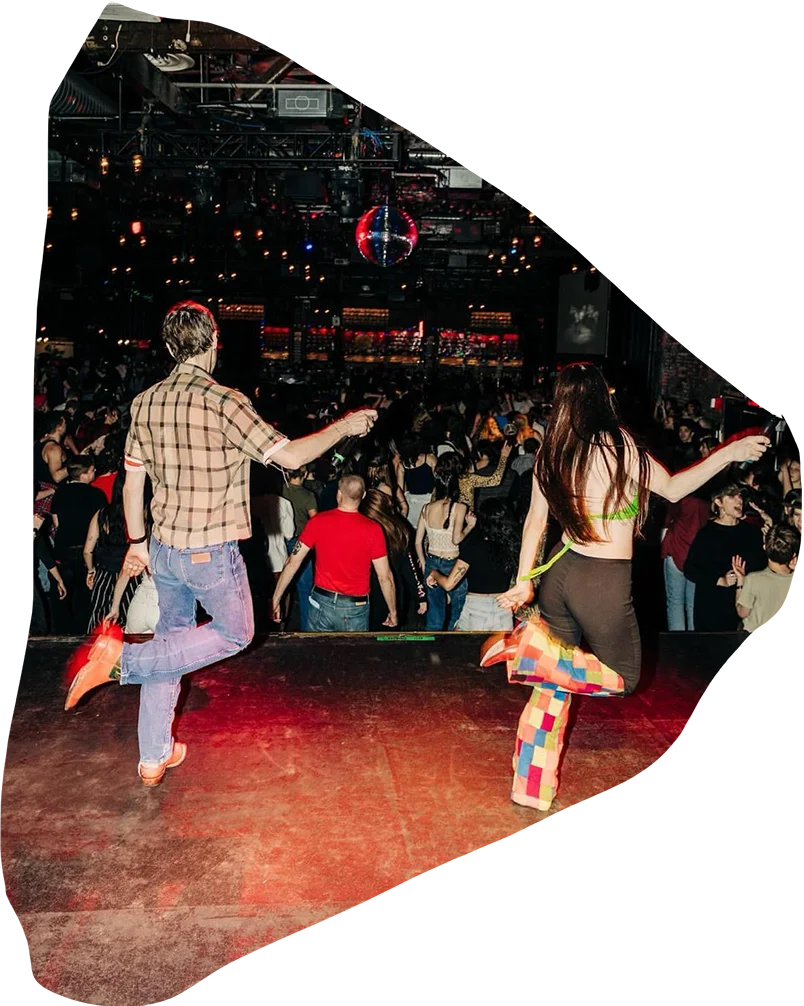 A lively scene of people line dancing in a dimly lit, vibrant club, with a person in the foreground wearing colorful pants and dancing energetically.
