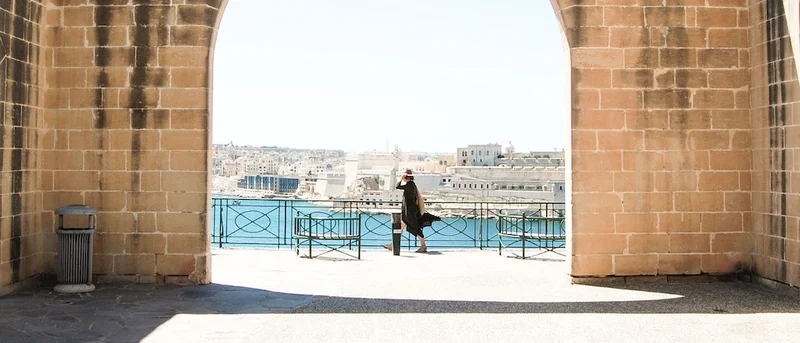 A view through a brick wall's arched opening. A person in a black dress walks past a body of water and white buildings.