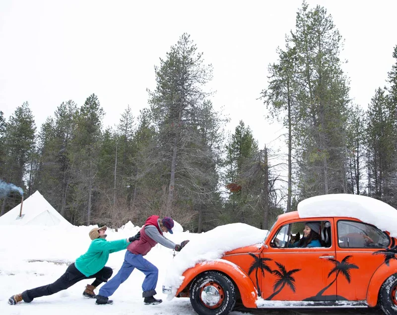 Two people pushing an orange VW Bug covered in snow.