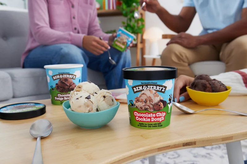 Coffee table with two pints of Ben & Jerry's ice cream in a living room setting. Two people are eating ice cream in the background.