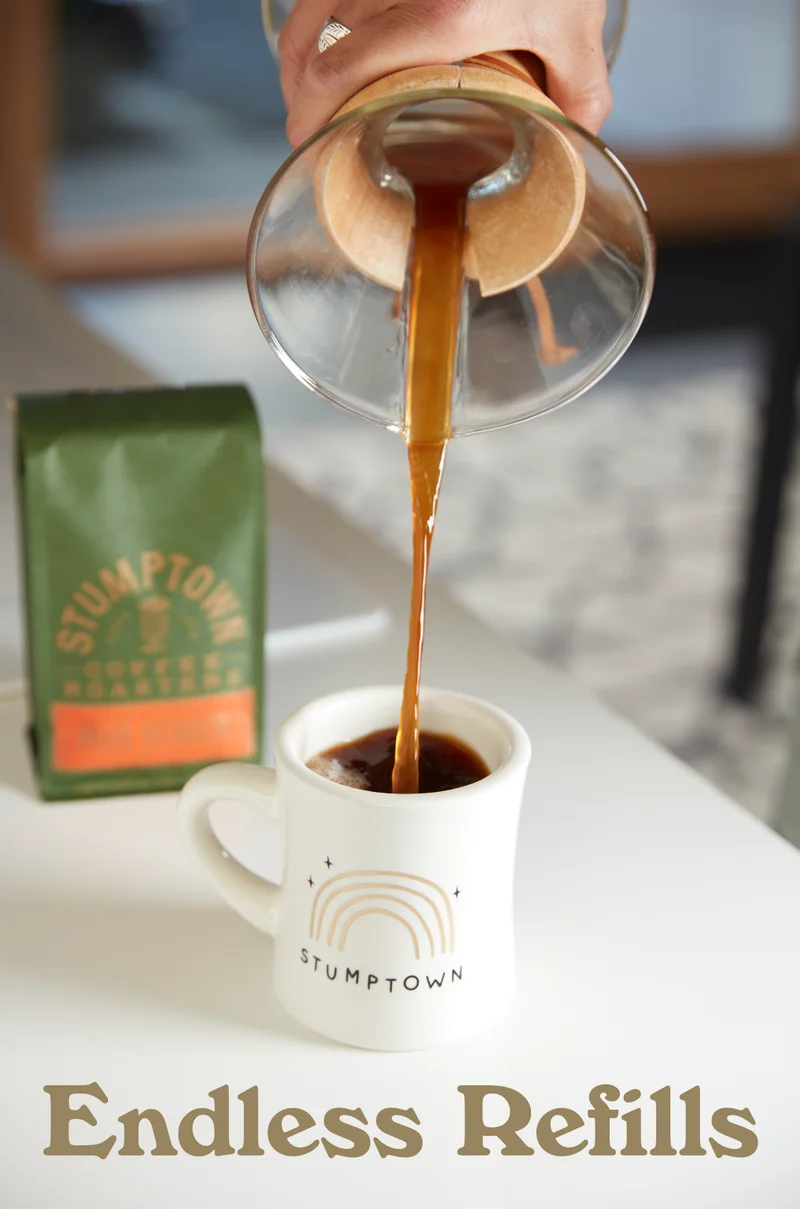 Hot coffee pouring from a Chemex into a Stumptown mug on a white table, with a bag of Hair Bender coffee in the background and the text “Endless Refills.”