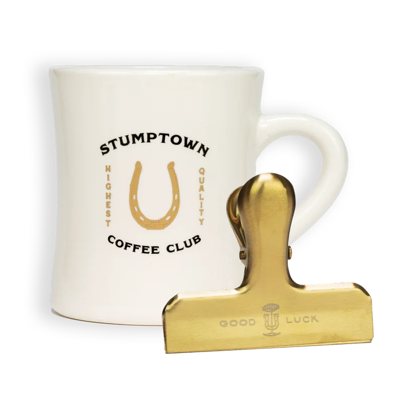 Stumptown Coffee Club ceramic diner mug in off-white with a gold horseshoe logo and “Highest Quality” text, placed behind a gold Stumptown bag clip engraved with “Good Luck”.