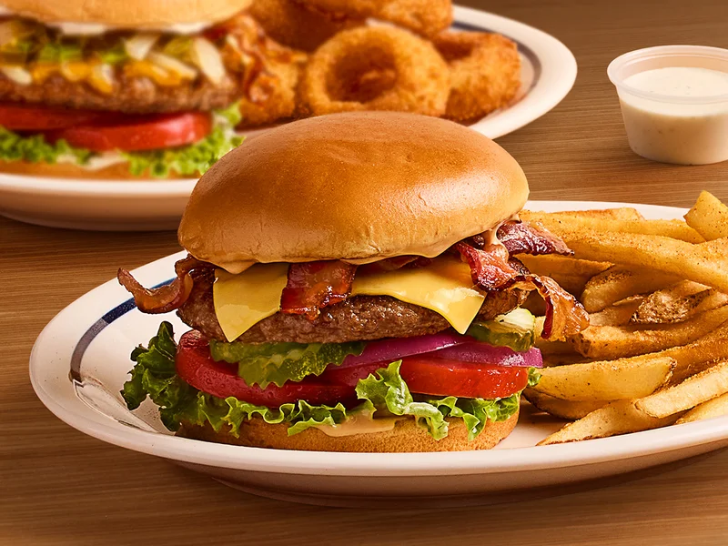 A classic IHOP Steakburger on a plate with fries, set on a table. A second Steakburger with a side of onion rings is on a plate in the background.