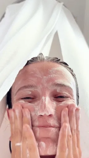 A person in the shower has suds all over their face, they rub their face and wiggle their eyebrows.