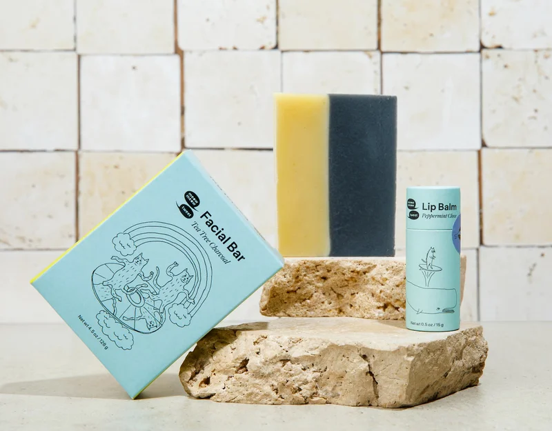 A blue soap box, a bar of soap, and a lip balm in a blue tube are arranged one and next to a couple slabs of marble, the Meow Meow Tweet Logo in the top left corner..