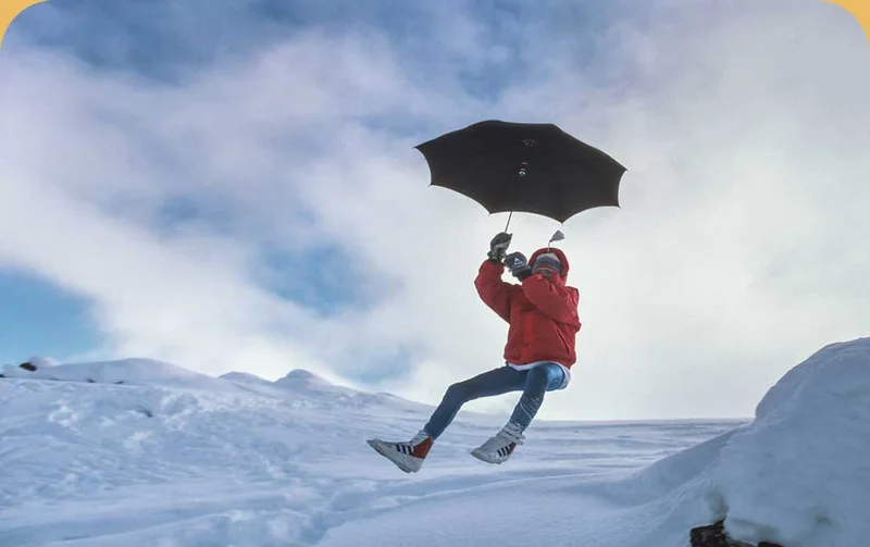 A person in a puffy jacket holding an umbrella jumps off a snow-covered hill.