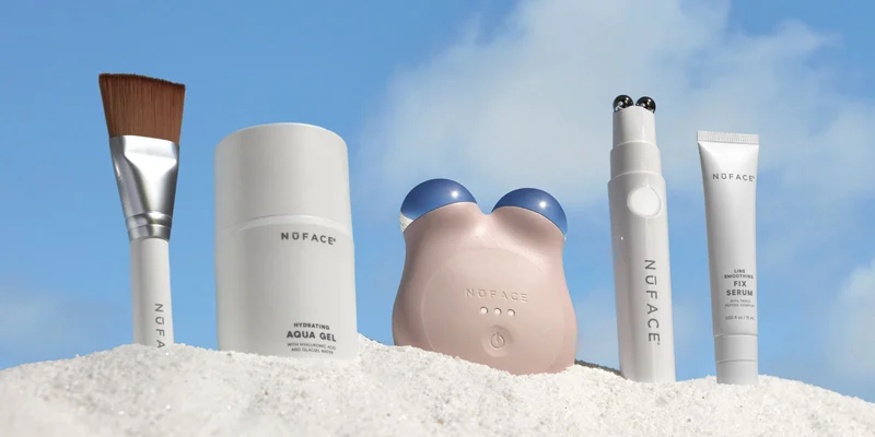 Clean Sweep Applicator Brush, Aqua Gel Microcurrent Activator, NuFACE MINI+ in Sandy Rose, NuFACE FIX in White, and NuFACE FIX Serum in the sand against a blue sky