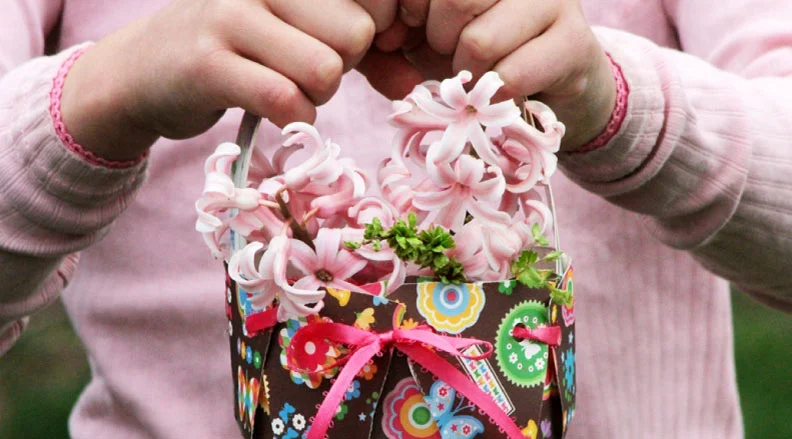 Paper Baskets: (Image of a child holding a DIY basket decorated with colorful paper and filled with pink flowers) Paper Baskets: (Image of a child holding a DIY basket decorated with colorful paper and filled with pink flowers)