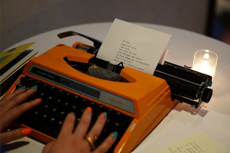 Two hands typing a poem on an orange typewriter