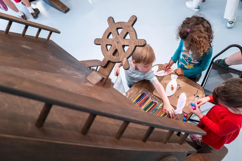 Three kids drawing with colored pencils beneath a ship replica