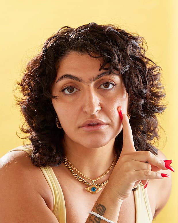 A person with brown eyes and brown curly hair applies cream to their cheek with a very long red nail