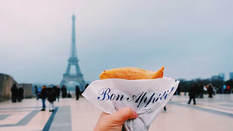 Image description: Person holding up crepe with view of Eiffel Tower in Paris in the background.