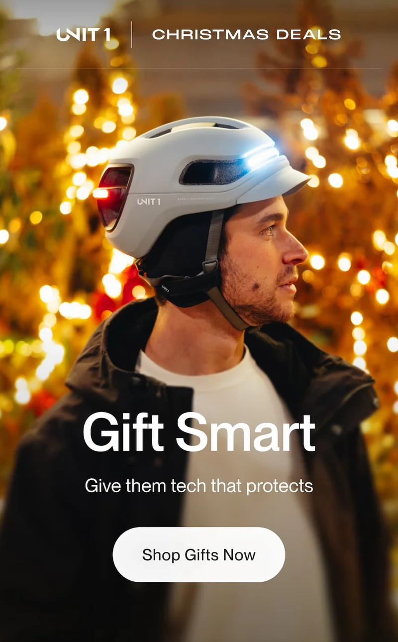 UNIT 1 smart helmet with integrated front and rear lights worn by a rider at night, Christmas lights in the background, with the message “Gift Smart – Give them tech that protects” and a “Shop Gifts Now” button.