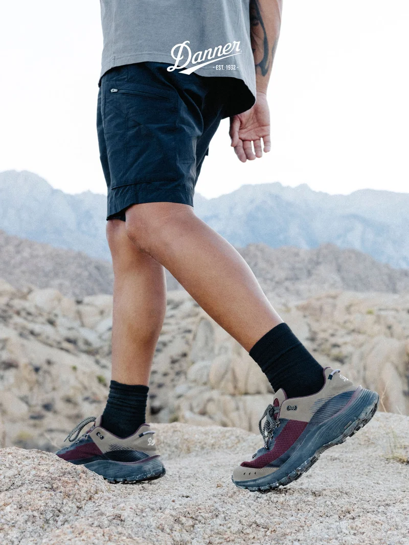 A hiker stands on a rocky desert landscape, wearing Trail Junction hiking shoes in Kangaroo/Chocolate. The Danner logo is overlaid at the top of the image.