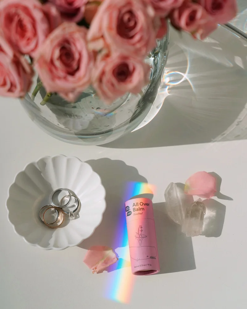 A pink tube lays on a table with a rainbow prism flashing over it. A vase of roses is in the foreground.