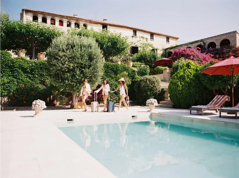 Three women in swimwear and cover-ups walk beside a pool with rolling suitcases at a Mediterranean-style villa surrounded by lush greenery and blooming flowers.