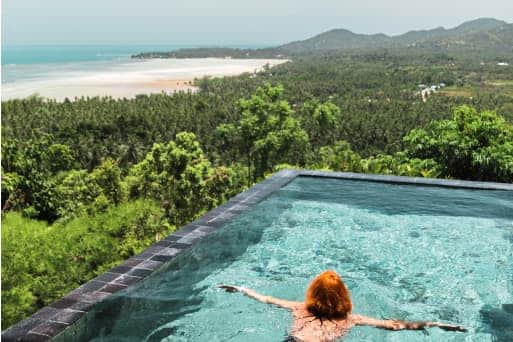 Person relaxing in an infinity pool overlooking a lush tropical forest and distant beach coastline.