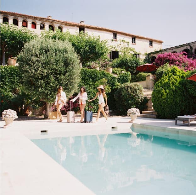 Three women in swimwear and cover-ups walk beside a pool with rolling suitcases at a Mediterranean-style villa surrounded by lush greenery and blooming flowers.