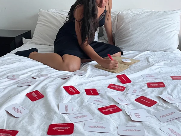 image of a women on her bed with cards from the We're Not Really Strangers card game spread out and she is journaling her answers.