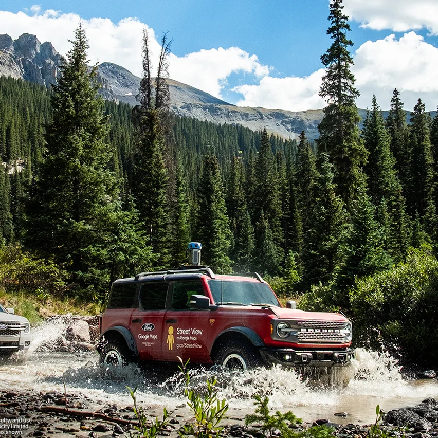 Red Ford Bronco SUV with Google Maps and Street View logos, splashing through a stream.