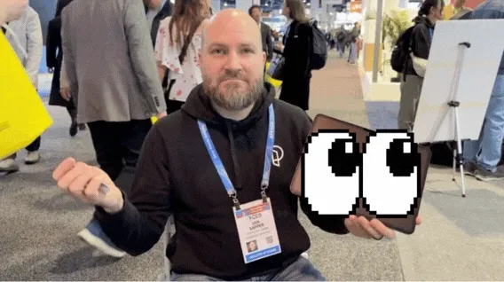 Image of Jan holding an iPad and shrugging his shoulders on a small chair in the middle of CES.