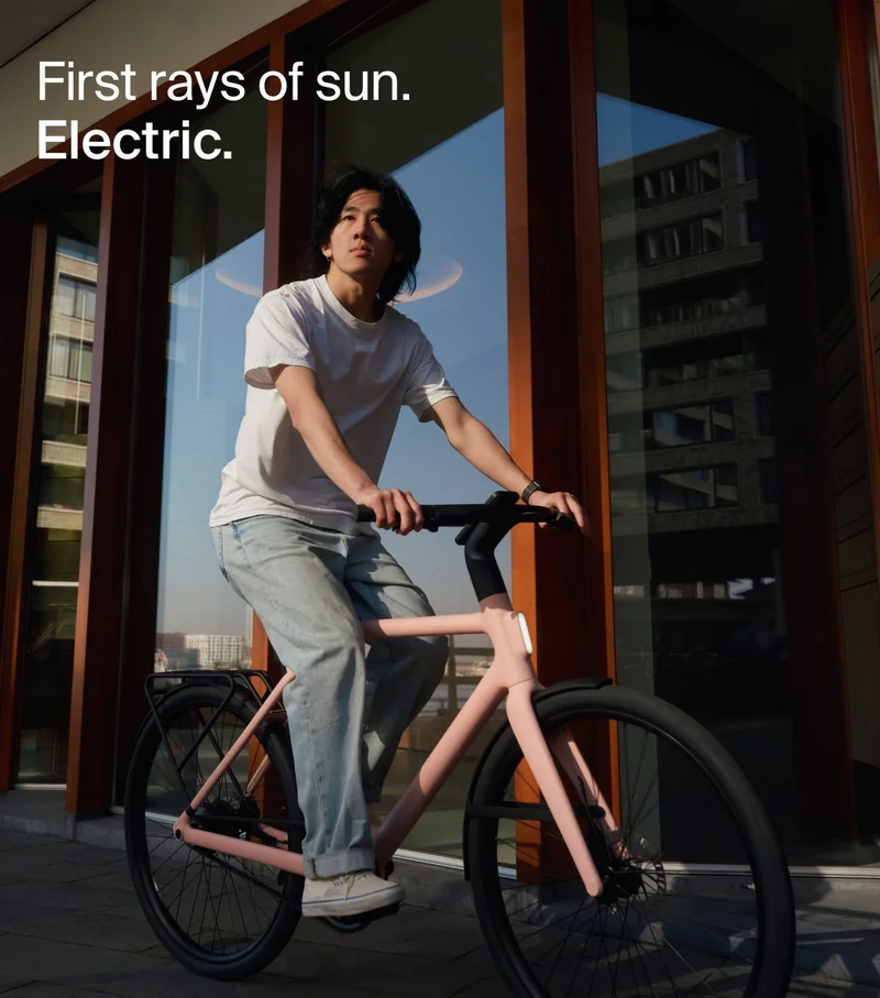 A person rides a Cowboy electric bike in the Dawn colorway, bathed in the morning light. The text overlay reads: “First rays of sun. Electric.”