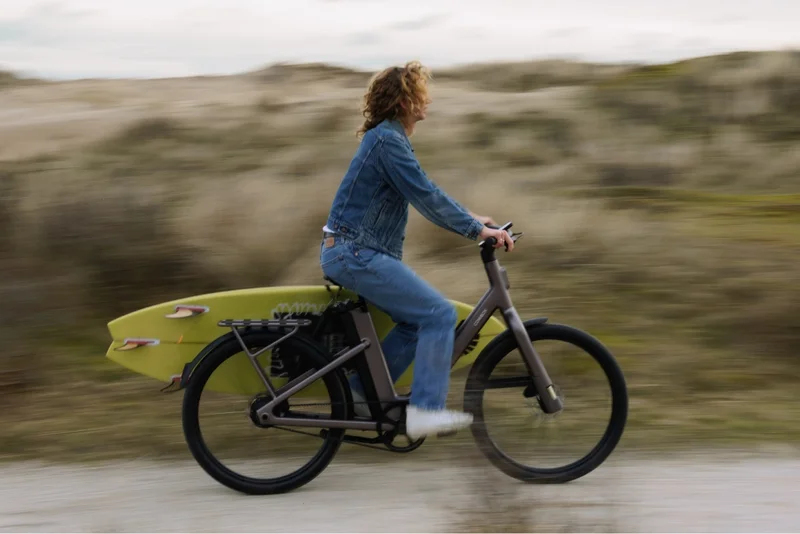 A group of three friends standing with their Cowboy bikes in a city setting. The person in the bright yellow jacket sits on a green bike, smiling, while the others lean casually against their bikes and chat.