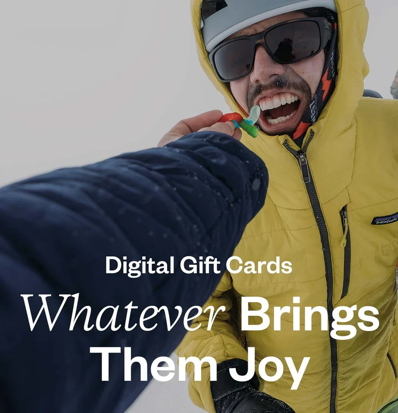 The outstretched arm of the photographer feeds a gummy bear to a person standing on a mountain summit in the winter.