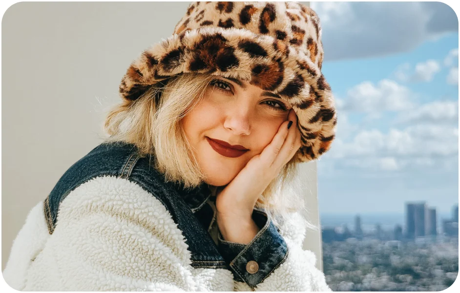 Well-lit image of a woman with a soft smile in a cheetah print hat.