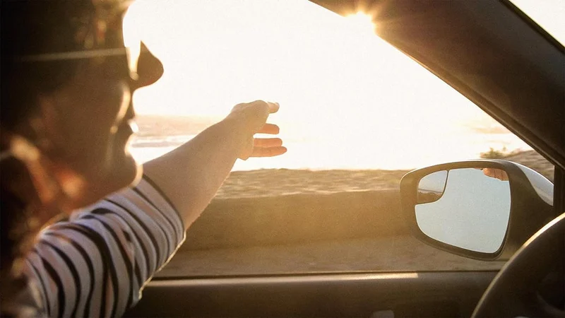Image description: Woman driving by herself with view of sunshine and beach.