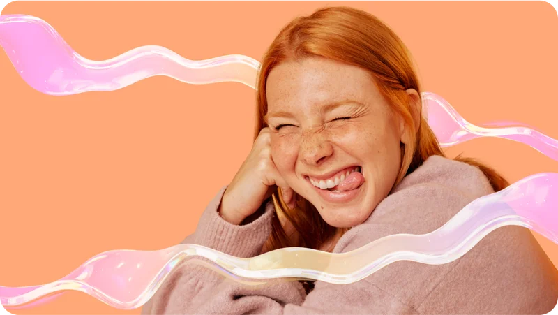 Smiling red-haired woman winking and sticking out her tongue, with iridescent light streaks on a warm peach background.