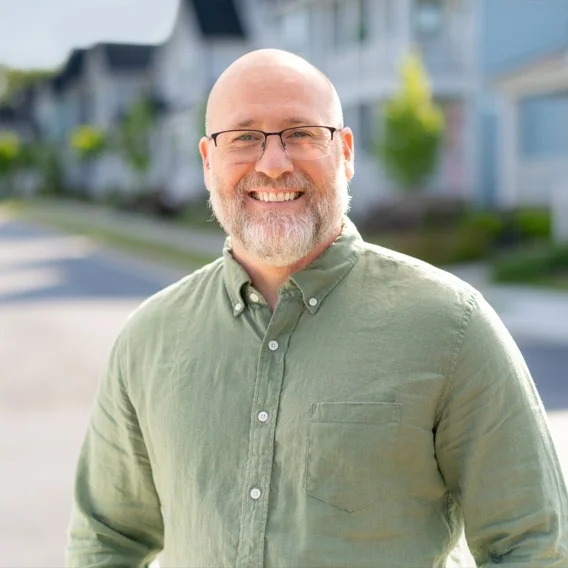 a man wearing glasses smiling at the camera on a suburban street